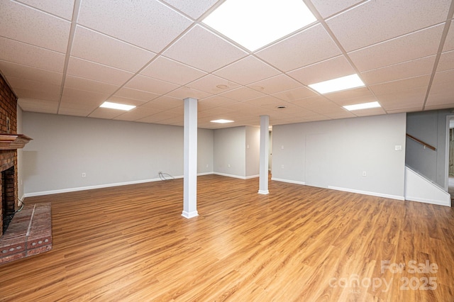 basement with a drop ceiling, a brick fireplace, and light hardwood / wood-style flooring