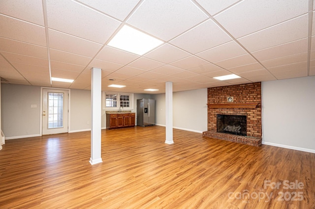 basement with stainless steel refrigerator with ice dispenser, a drop ceiling, a brick fireplace, and hardwood / wood-style flooring