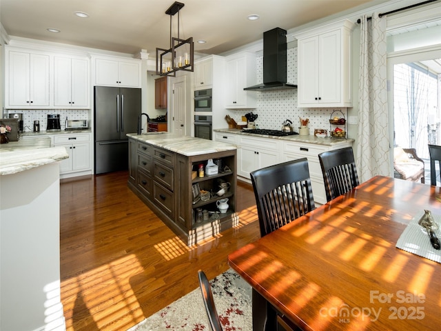 kitchen with appliances with stainless steel finishes, pendant lighting, white cabinetry, a kitchen island with sink, and wall chimney range hood