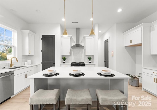 kitchen with wall chimney range hood, sink, dishwasher, white cabinetry, and decorative light fixtures