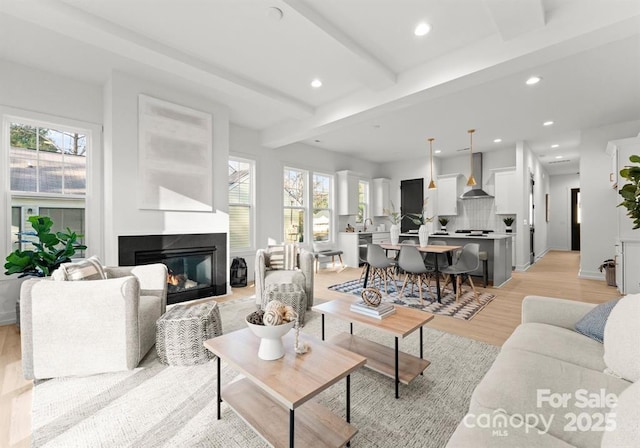 living area featuring plenty of natural light, recessed lighting, light wood-type flooring, beamed ceiling, and a glass covered fireplace