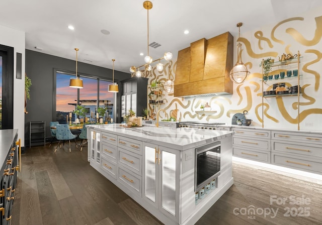 kitchen with white cabinetry, light stone counters, a center island with sink, decorative light fixtures, and custom exhaust hood