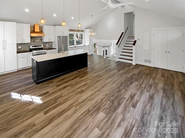 kitchen with wall chimney exhaust hood, hanging light fixtures, appliances with stainless steel finishes, a kitchen island with sink, and white cabinets