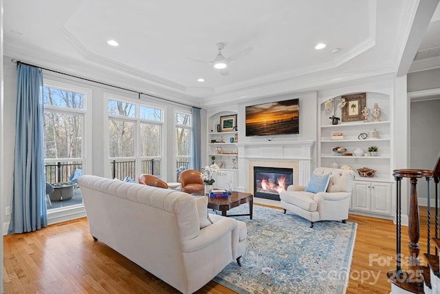 living area featuring built in features, a ceiling fan, a glass covered fireplace, crown molding, and light wood-style floors