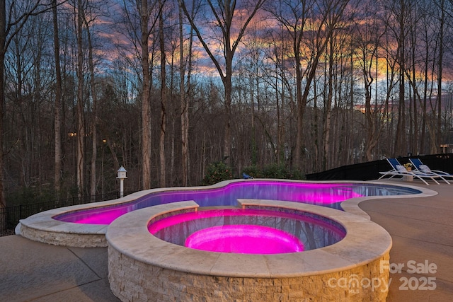 pool at dusk featuring fence, an outdoor pool, a patio, and an in ground hot tub