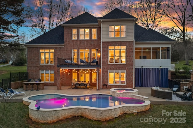 back of property at dusk featuring a patio, a balcony, an in ground hot tub, brick siding, and an outdoor living space with a fire pit