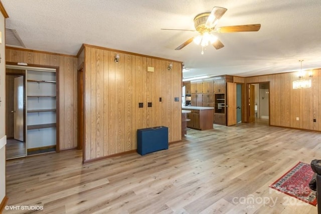 unfurnished living room with a textured ceiling, light wood-type flooring, ornamental molding, wooden walls, and ceiling fan with notable chandelier