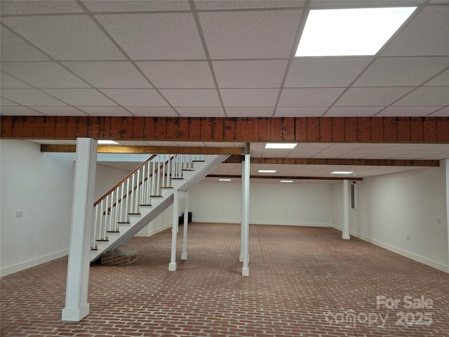 finished below grade area featuring stairway, baseboards, brick floor, and a drop ceiling