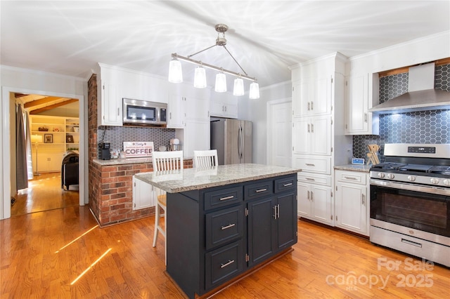 kitchen featuring appliances with stainless steel finishes, white cabinetry, a kitchen island, decorative light fixtures, and wall chimney exhaust hood