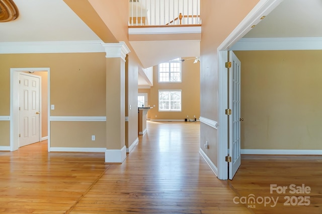 hall with ornate columns, a towering ceiling, ornamental molding, wood finished floors, and baseboards