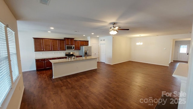 kitchen with appliances with stainless steel finishes, a kitchen island with sink, light stone countertops, dark hardwood / wood-style flooring, and ceiling fan with notable chandelier