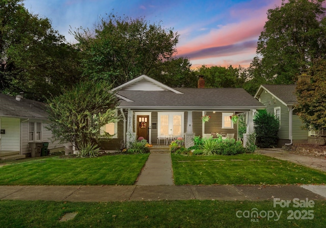 view of front facade with a front yard, covered porch, roof with shingles, and a chimney