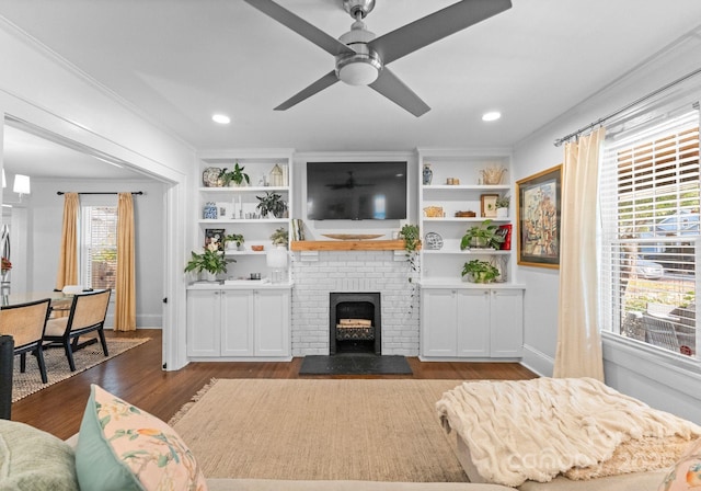 living area featuring dark wood-type flooring, a ceiling fan, built in features, a brick fireplace, and crown molding