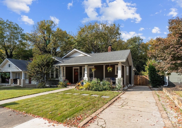 bungalow featuring a chimney, covered porch, concrete driveway, fence, and a front lawn