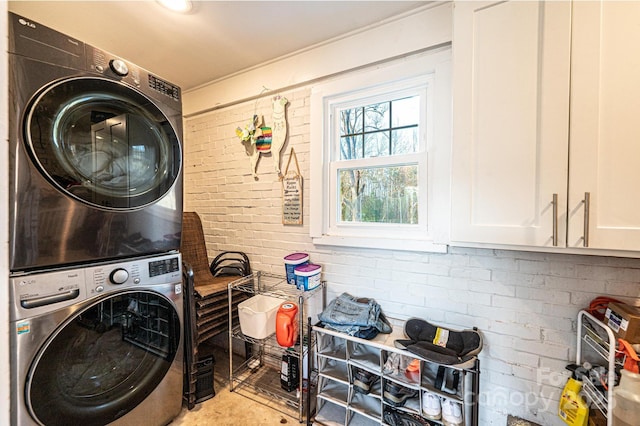 clothes washing area with stacked washer / dryer, cabinet space, and brick wall