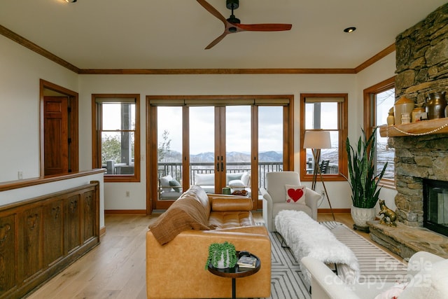 living room with a stone fireplace, ceiling fan, crown molding, a mountain view, and light wood-type flooring