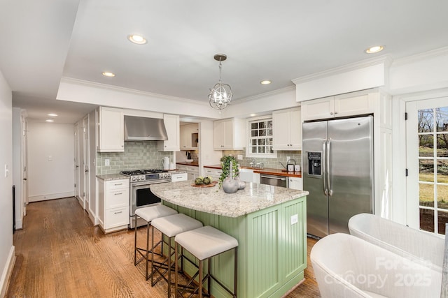 kitchen with stainless steel appliances, a kitchen island, white cabinetry, hanging light fixtures, and wall chimney exhaust hood