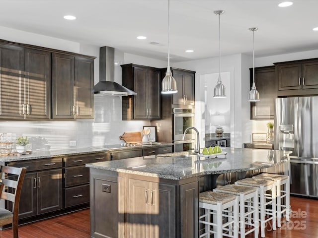 kitchen featuring wall chimney range hood, stone counters, appliances with stainless steel finishes, hanging light fixtures, and a center island with sink