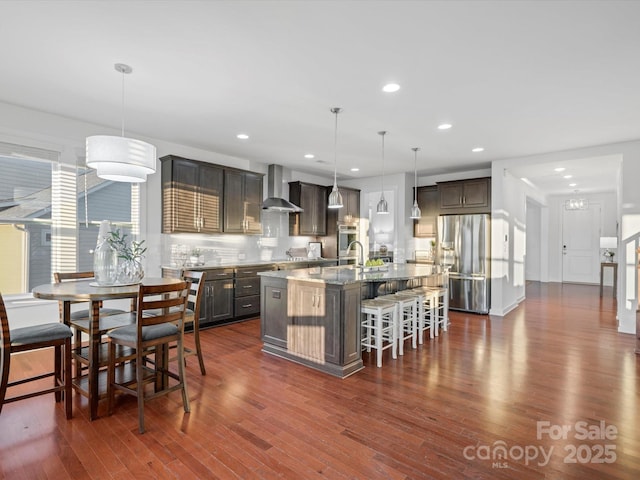 kitchen with wall chimney exhaust hood, a breakfast bar, hanging light fixtures, appliances with stainless steel finishes, and an island with sink