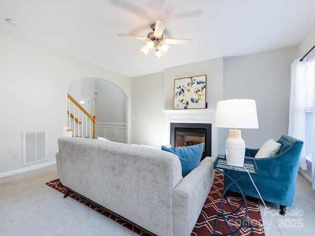 carpeted living area with stairway, visible vents, a fireplace, arched walkways, and ceiling fan