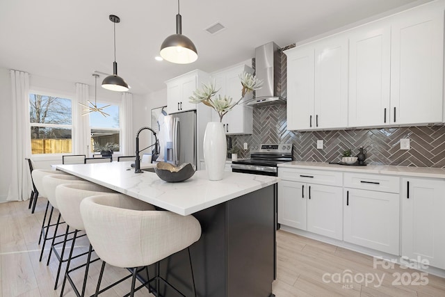 kitchen with decorative light fixtures, wall chimney range hood, stainless steel appliances, a kitchen island with sink, and white cabinets