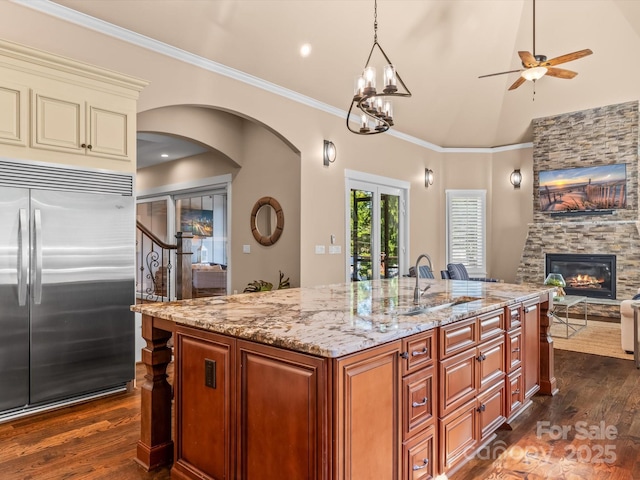 kitchen featuring built in fridge, a stone fireplace, decorative light fixtures, an island with sink, and crown molding