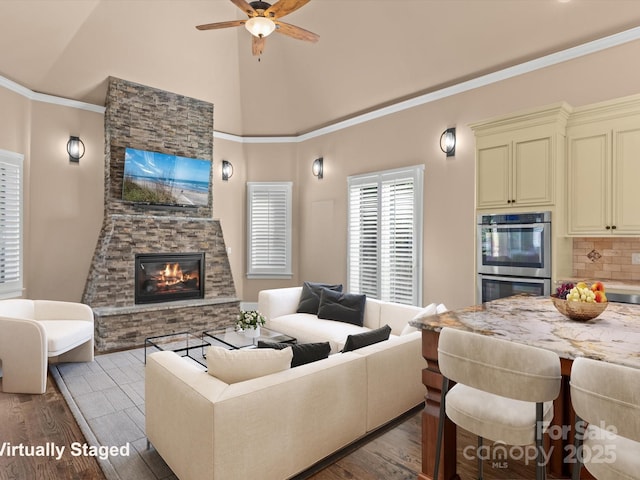 living room with dark wood-type flooring, ornamental molding, ceiling fan, a stone fireplace, and high vaulted ceiling