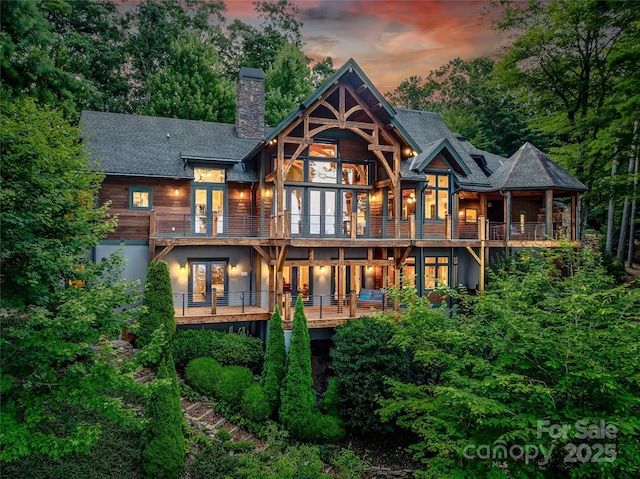 back of house at dusk with roof with shingles, french doors, a chimney, and a balcony