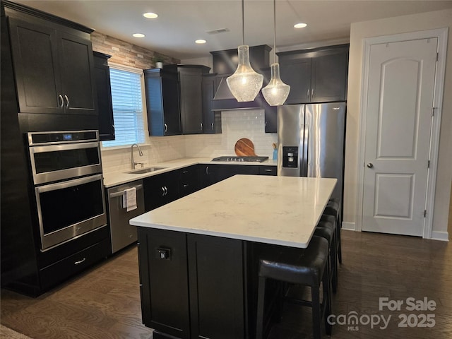 kitchen with appliances with stainless steel finishes, dark cabinets, a center island, wall chimney range hood, and a sink