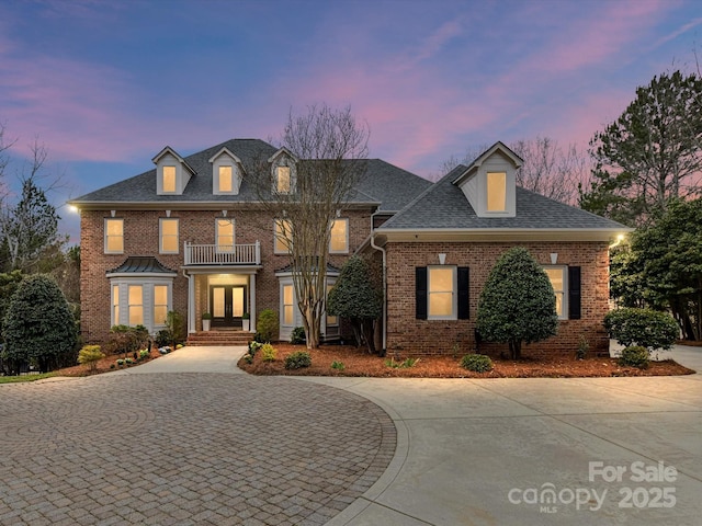 colonial house featuring brick siding, curved driveway, a balcony, and a shingled roof