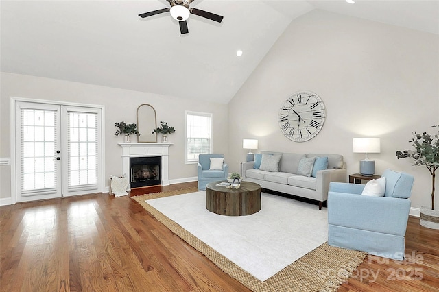 living area featuring a fireplace, a ceiling fan, wood finished floors, high vaulted ceiling, and baseboards