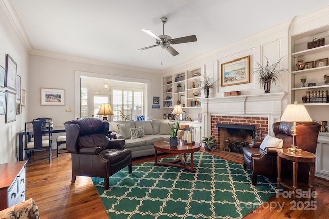 living room with built in shelves, crown molding, wood-type flooring, ceiling fan, and a fireplace