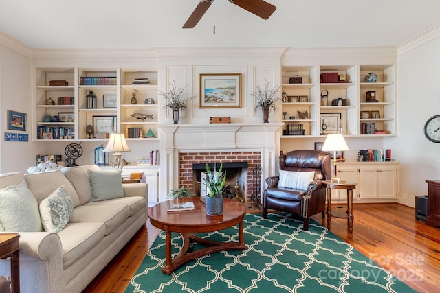 living area featuring crown molding, a brick fireplace, built in features, hardwood / wood-style flooring, and ceiling fan