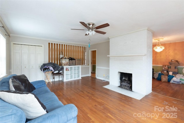 living area with ceiling fan with notable chandelier, ornamental molding, a brick fireplace, and wood finished floors