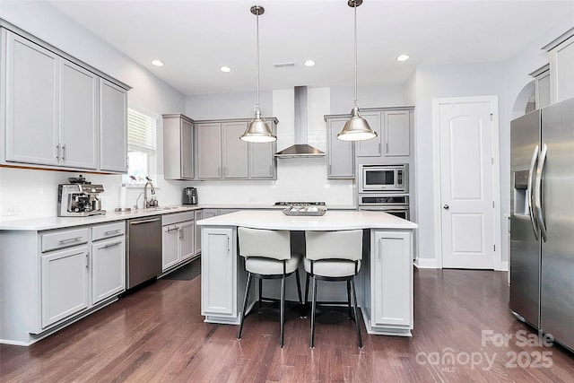 kitchen featuring wall chimney range hood, appliances with stainless steel finishes, gray cabinetry, a center island, and decorative light fixtures