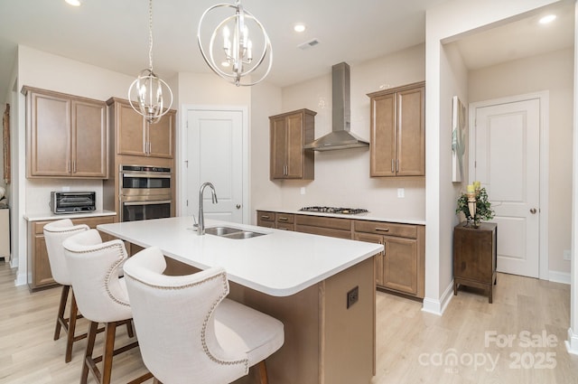 kitchen with wall chimney exhaust hood, sink, appliances with stainless steel finishes, pendant lighting, and a kitchen island with sink