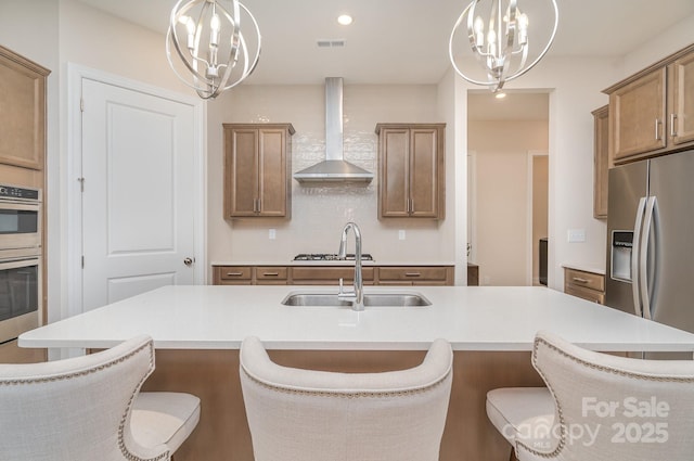 kitchen featuring appliances with stainless steel finishes, wall chimney exhaust hood, a center island with sink, and a notable chandelier