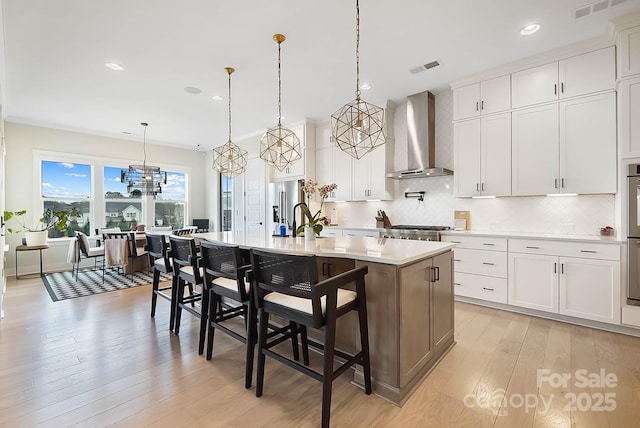 kitchen featuring pendant lighting, white cabinets, a kitchen breakfast bar, a kitchen island with sink, and wall chimney range hood