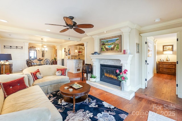 living room with ornate columns, wood-type flooring, ceiling fan with notable chandelier, and crown molding