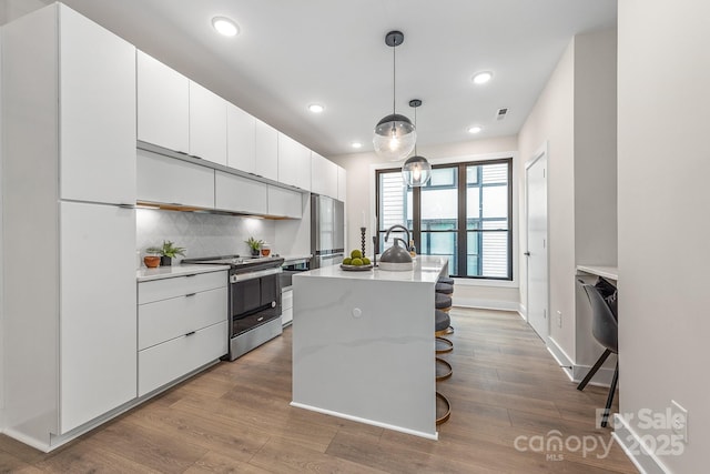 kitchen featuring stainless steel range with electric stovetop, white cabinetry, a kitchen island with sink, a kitchen breakfast bar, and decorative light fixtures