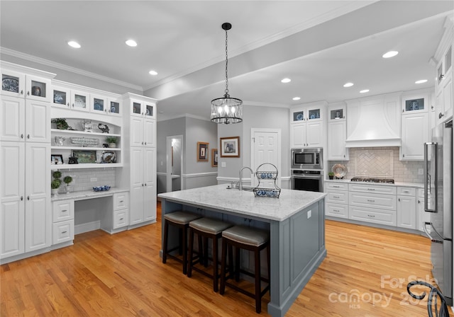 kitchen with appliances with stainless steel finishes, premium range hood, built in desk, white cabinets, and a center island with sink