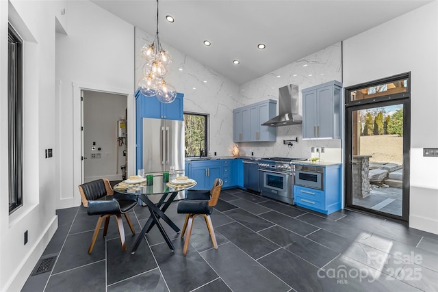 kitchen featuring blue cabinetry, premium appliances, decorative light fixtures, and wall chimney range hood