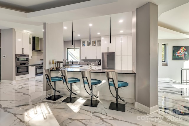 kitchen with white cabinetry, wall chimney range hood, stainless steel appliances, and hanging light fixtures