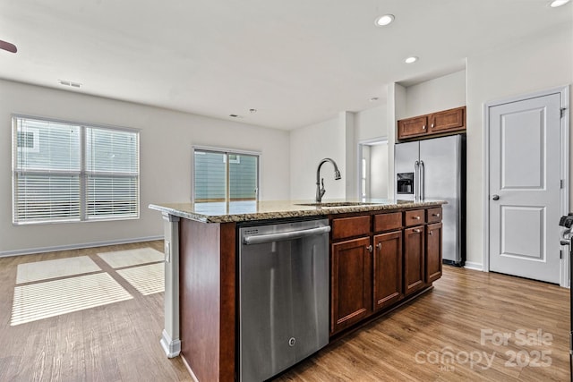 kitchen with sink, a kitchen island with sink, stainless steel appliances, light stone countertops, and light wood-type flooring