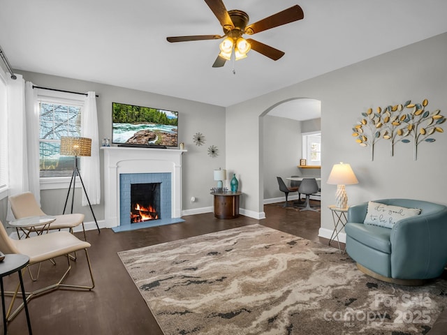 living room with baseboards, arched walkways, dark wood-style floors, ceiling fan, and a brick fireplace