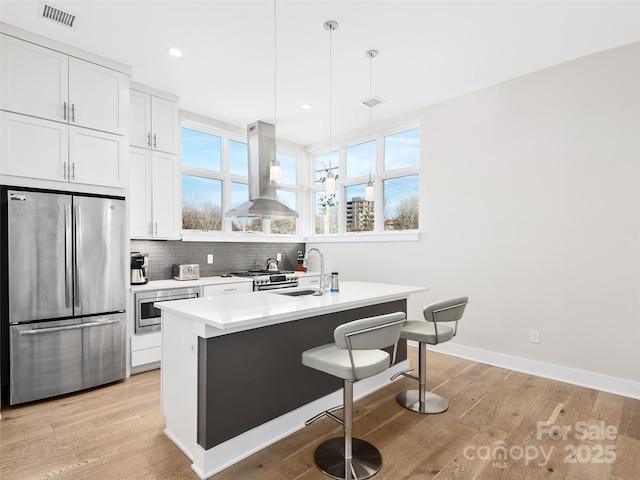 kitchen with appliances with stainless steel finishes, island range hood, white cabinets, hanging light fixtures, and a center island with sink