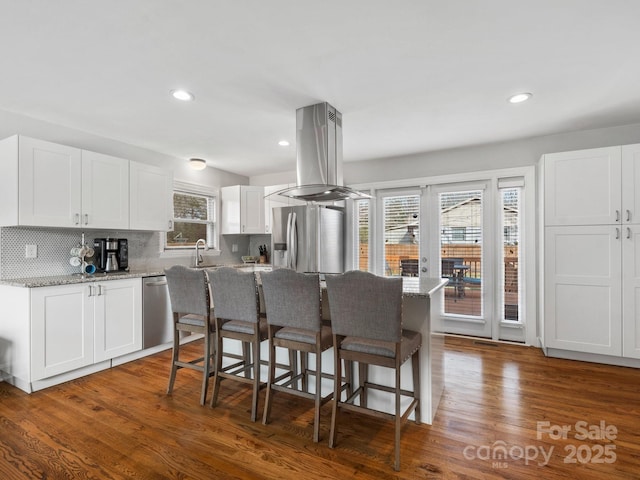 kitchen with island range hood, stainless steel appliances, white cabinets, light stone countertops, and dark wood-style floors