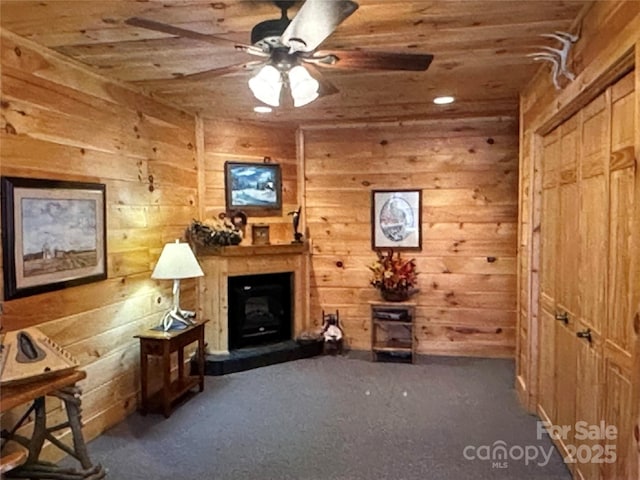 sitting room featuring wooden ceiling, wooden walls, a fireplace with raised hearth, and a ceiling fan