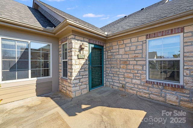 property entrance with stone siding, roof with shingles, and a patio area