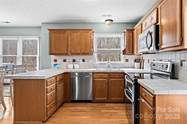 kitchen with tile countertops, sink, kitchen peninsula, stainless steel appliances, and light hardwood / wood-style flooring